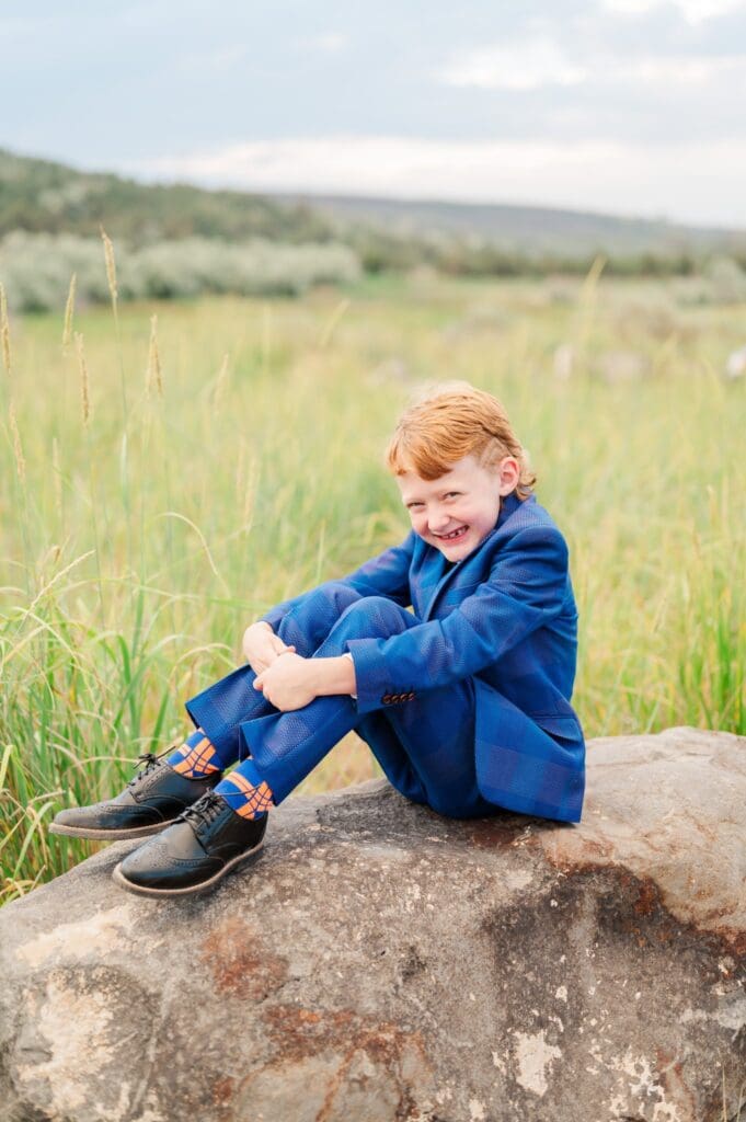 Child sitting on a large rock while wearing in navy blue suit grinning during LDS baptism session in southeast Idaho